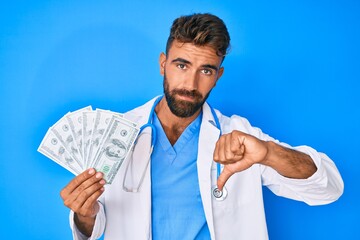 Young hispanic man wearing doctor uniform holding dollars with angry face, negative sign showing...