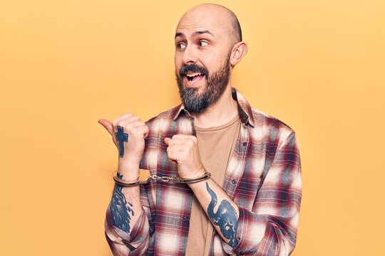 Young handsome man wearing prisoner handcuffs pointing thumb up to the side smiling happy with open mouth
