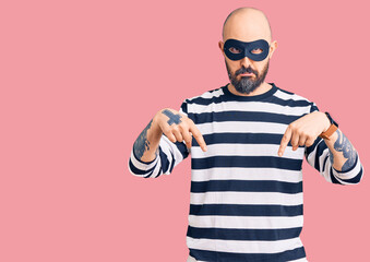 Young handsome man wearing burglar mask pointing down looking sad and upset, indicating direction with fingers, unhappy and depressed.