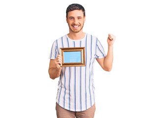 Handsome young man with bear holding empty frame screaming proud, celebrating victory and success very excited with raised arms