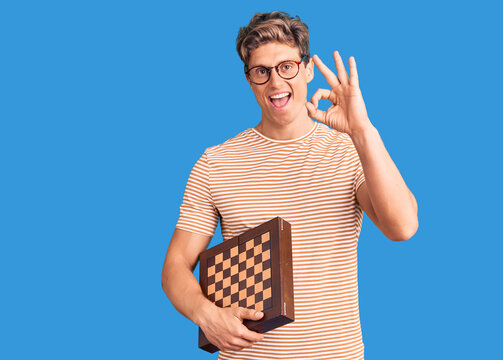 Young handsome man holding chess doing ok sign with fingers, smiling friendly gesturing excellent symbol