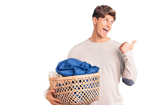 Young handsome man holding laundry basket pointing thumb up to the side smiling happy with open mouth
