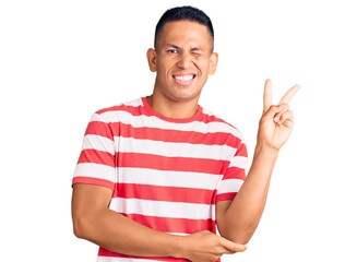 Young handsome latin man wearing casual clothes smiling with happy face winking at the camera doing victory sign. number two.