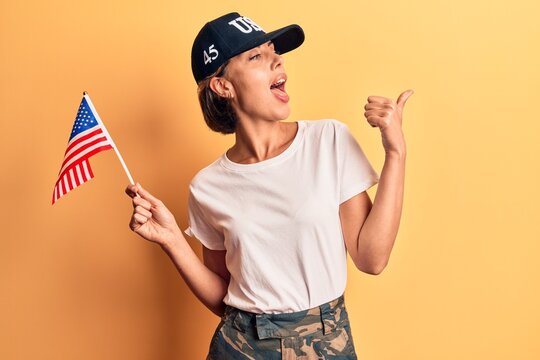 Young beautiful woman wearing usa cap holding united states flag pointing thumb up to the side smiling happy with open mouth