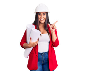 Young beautiful brunette woman wearing hardhat holding architect blueprint smiling happy pointing with hand and finger to the side
