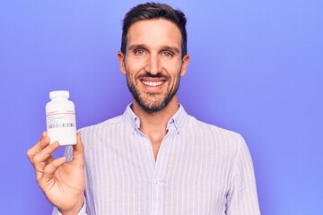 Young handsome man holding jar of medicine pills standing over isolated purple background looking positive and happy standing and smiling with a confident smile showing teeth