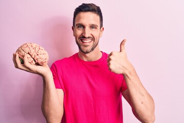 Young handsome man asking for care memory holding brain over isolated pink background smiling happy...