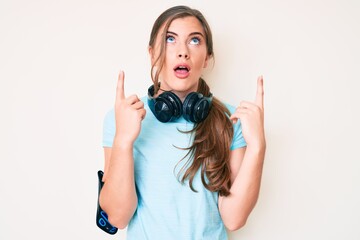 Fototapeta premium Beautiful young caucasian woman wearing gym clothes and using headphones amazed and surprised looking up and pointing with fingers and raised arms.