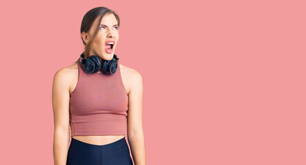 Obraz premium Beautiful caucasian young woman wearing gym clothes and using headphones angry and mad screaming frustrated and furious, shouting with anger. rage and aggressive concept.
