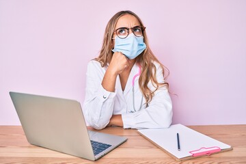 Young beautiful blonde woman wearing doctor uniform and medical mask serious face thinking about...