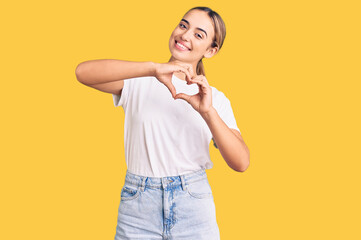 Young beautiful blonde woman wearing casual white tshirt smiling in love doing heart symbol shape with hands. romantic concept.