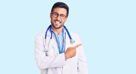 Fototapete Artz Junger hispanischer Mann, der Arztuniform und Stethoskop trägt, fröhlich mit einem Lächeln im Gesicht, das mit Hand und Finger mit glücklichem und natürlichem Ausdruck zur Seite zeigt