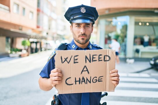 Young handsome hispanic policeman wearing police uniform with serious expression holding we need a change banner at town street