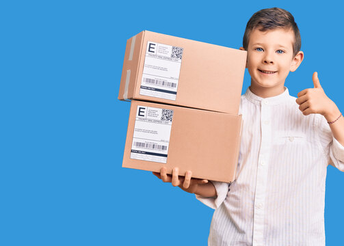 Cute blond kid holding delivery package smiling happy and positive, thumb up doing excellent and approval sign