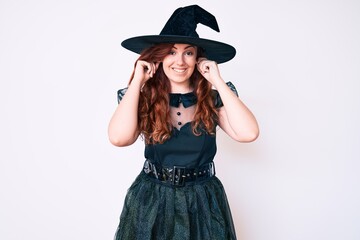 Young beautiful woman wearing witch halloween costume smiling pulling ears with fingers, funny gesture. audition problem