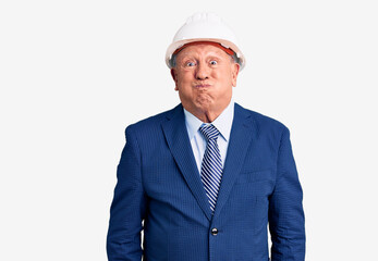 Senior handsome grey-haired man wearing suit and architect hardhat puffing cheeks with funny face. mouth inflated with air, crazy expression.