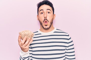 Young hispanic man holding chickpeas bowl scared and amazed with open mouth for surprise, disbelief face