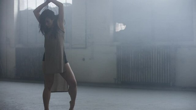 Beautiful dancer dances, stops abruptly then dances passionately with lots of energy surrounded by fog and mist in industrial warehouse