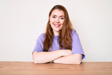 Young redhead woman wearing casual clothes sitting on the table happy face smiling with crossed arms looking at the camera. positive person.