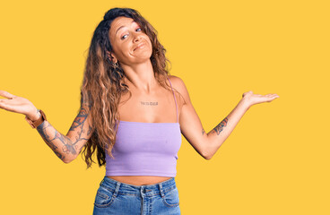 Young hispanic woman with tattoo wearing casual clothes clueless and confused expression with arms...