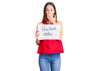 Beautiful brunette young woman holding positive vibes banner covering mouth with hand, shocked and afraid for mistake. surprised expression