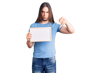 Fototapeta premium Young adult man with long hair holding empty white chalkboard with angry face, negative sign showing dislike with thumbs down, rejection concept