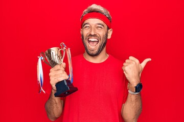 Young handsome blond sportsman with beard winning trophy over isolated red background pointing thumb up to the side smiling happy with open mouth