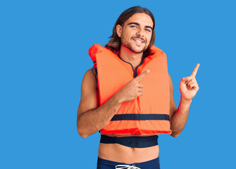 Young handsome man wearing nautical lifejacket smiling and looking at the camera pointing with two...