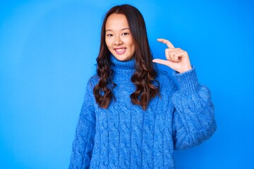 Young beautiful chinese girl wearing casual winter sweater smiling and confident gesturing with hand doing small size sign with fingers looking and the camera. measure concept.