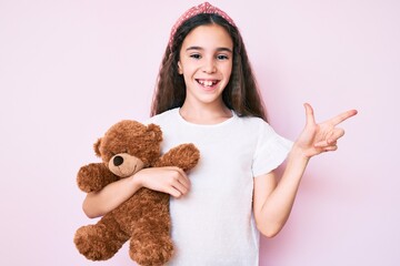 Cute hispanic child girl holding teddy bear smiling happy pointing with hand and finger to the side