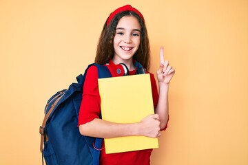 Cute hispanic child girl wearing student backpack and headphones holding book surprised with an idea or question pointing finger with happy face, number one