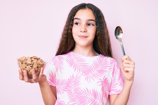 Cute hispanic girl holding cornflakes and spoon smiling looking to the side and staring away thinking.
