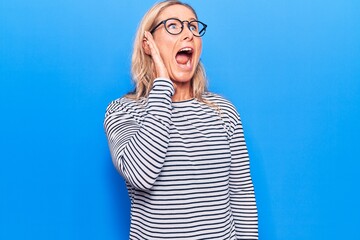 Plakat Middle age caucasian blonde woman wearing casual striped sweater and glasses shouting and screaming loud to side with hand on mouth. communication concept.