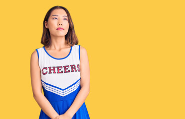 Young beautiful chinese girl wearing cheerleader uniform smiling looking to the side and staring...