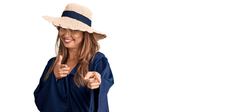 Middle age hispanic woman wearing summer hat pointing fingers to camera with happy and funny face. good energy and vibes.