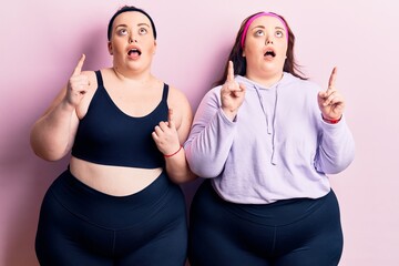 Young plus size twins wearing sportswear amazed and surprised looking up and pointing with fingers and raised arms.