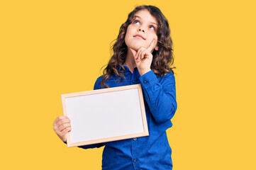 Cute hispanic child with long hair holding empty white chalkboard serious face thinking about...