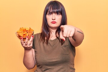 Young plus size woman holding nachos potato chips pointing with finger to the camera and to you, confident gesture looking serious