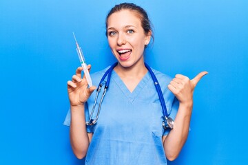 Young blonde woman wearing doctor uniform holding syringe pointing thumb up to the side smiling happy with open mouth