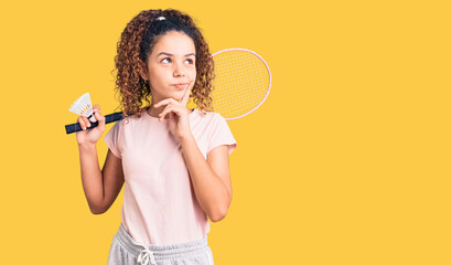 Beautiful kid girl with curly hair holding badminton racket and shuttlecock serious face thinking about question with hand on chin, thoughtful about confusing idea - obrazy, fototapety, plakaty