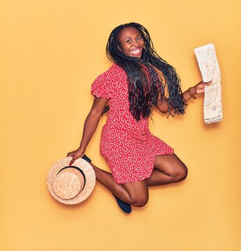 Young beautiful african american woman wearing summer clothes smiling happy. Jumping with smile on face holding city map and hat over isolated yellow background.