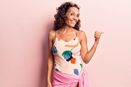 Middle age beautiful woman wearing swimwear and sunglasses smiling with happy face looking and pointing to the side with thumb up.