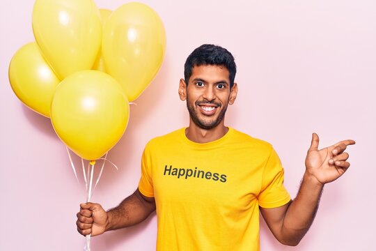 Young latin man wearing t shirt with happiness message holding balloons smiling happy pointing with hand and finger to the side