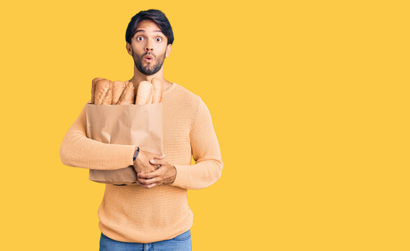Handsome hispanic man holding paper bag with bread scared and amazed with open mouth for surprise, disbelief face