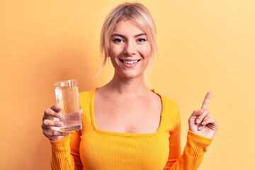 Young beautiful blonde woman drinking glass of water over isolated yellow background smiling happy pointing with hand and finger to the side