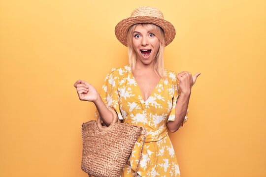 Young beautiful blonde woman on vacation wearing summer hat and dress holding wicker bag pointing thumb up to the side smiling happy with open mouth