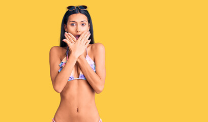 Young beautiful latin girl wearing bikini and sunglasses shocked covering mouth with hands for...