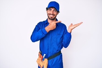 Handsome young man with curly hair and bear weaing handyman uniform amazed and smiling to the camera while presenting with hand and pointing with finger.