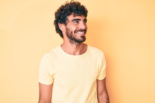 Handsome young man with curly hair and bear wearing casual tshirt over yellow background looking to side, relax profile pose with natural face and confident smile.