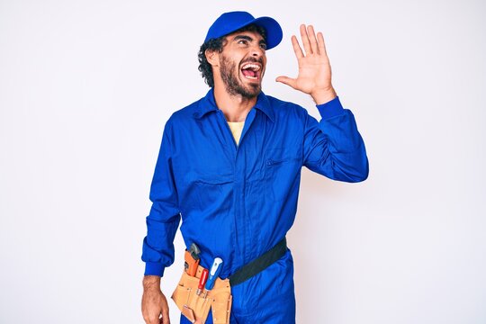 Handsome young man with curly hair and bear weaing handyman uniform shouting and screaming loud to side with hand on mouth. communication concept.